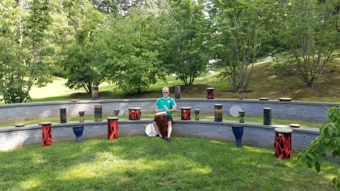 Drumming instructor seated on outdoor bench surrounded by a dozen drums.